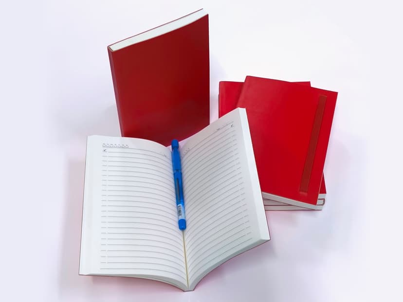 Stack of red rexine diaries with one opened to show ruled pages and a blue pen.